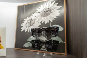 A pair of sunglasses is displayed on a clear stand in front of a framed picture of sunflowers.