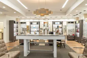 An interior view of an optical shop with a woman sitting at a desk, surrounded by displays of eyeglasses and other items on shelves, with a chandelier hanging from the ceiling, and chairs and a carpeted floor.
