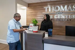 An adult man and woman are conversing near a wooden reception desk at Thomas Eye Center Optometry. The man is wearing glasses and a blue polo shirt, while the woman has long hair and is dressed in black. There is a potted plant and a sign on the desk, and the wall behind them has a wooden texture and is illuminated by a bright light. The room has a clean and professional atmosphere.