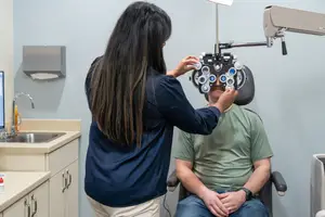 A woman is adjusting an ophthalmoscope on a man sitting in a chair in a medical office.