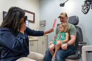 An optometrist examines a young girl's eyes while her father holds her steady, with a red and white object nearby.