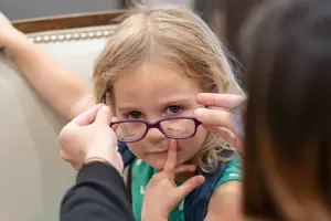 A young girl wearing glasses with a purple frame is having her glasses adjusted by a person in a black shirt.