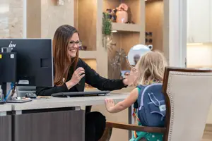 A woman is smiling and talking to a child while holding a mirror, while the child is holding the woman's hand. Behind them is a wooden cabinet with shelves and a monitor on a desk.