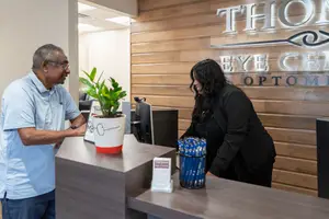 An older man and a woman are standing near a desk with a plant and a sign reading Thion Eye Center Optometry
