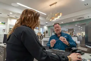 A man and woman are in an office, the man is wearing glasses and holding a pair of glasses while the woman is looking at him and working on a computer.
