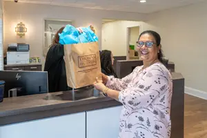 A smiling woman holds a gift bag at a reception desk with a printer and monitor in the background.