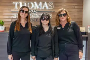 Three women in black clothing standing in front of a wooden wall with a sign reading Thomas Eye Center Homet