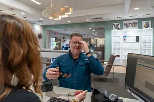 A man is sitting in front of a desk with a computer monitor, and he is holding a pair of sunglasses. A woman is standing next to him, and she is looking at the monitor. Behind them is a wall with a lamp and a picture frame. There are also chairs, a table, and a man standing near a window. There is also a glass wall with a picture of a dog on it.