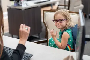 A young girl wearing glasses is sitting on a chair in an office with a woman wearing a ring and a desk with a computer monitor and keyboard