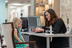 A woman is examining a young girl's eyes in a medical office with a computer on the desk