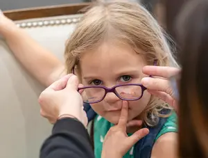 a little girl with blonde hair wearing purple glasses is looking at someone who is adjusting her glasses