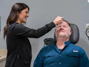 A woman in a black long-sleeve shirt is adjusting the glasses of a man in a blue shirt in a clinic setting.