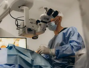 An eye doctor is performing a surgery with an ophthalmoscope while wearing a surgical mask and blue gown.
