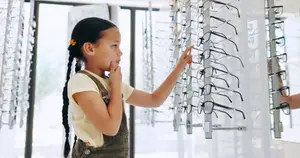 A girl is standing in front of a display of eyeglasses in a store, looking at them with her hand on her chin.