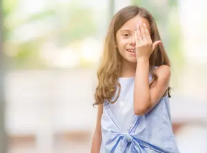 A young girl with brown hair covering her eyes with her hand and smiling while standing in front of a blurry background