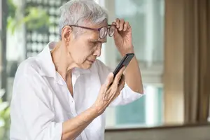 An older woman adjusts her glasses while looking at her phone in a room with a window and plants outside