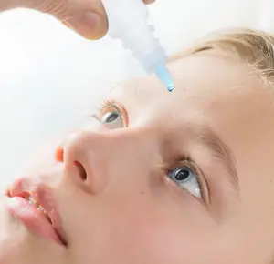A young girl is looking up as a person's hand administers eye drops to her left eye