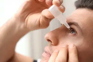 A person with a blurry face and hand is applying eye drops on his left eye. He is holding a white bottle with a dropper in his right hand, and his right hand is on his eye. He is looking down with a blurry face. He is wearing a black shirt and has a black beard and black hair.