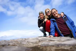 Three people are smiling and posing for a photo on a rocky cliff under a blue sky with white clouds.