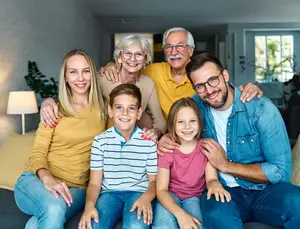 A family of four, including two grandparents, a mother, a father, a boy, and a girl, are sitting on a couch and smiling for a photo.