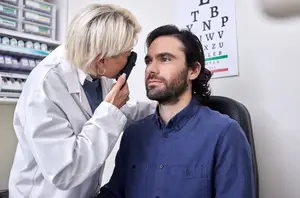 A woman optometrist is examining a man's eyes using an ophthalmoscope in a medical office.