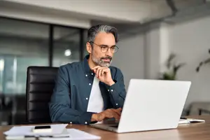 A man with gray hair wearing glasses and a blue shirt is working on his laptop while sitting at a desk in an office.