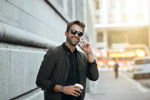 A man wearing sunglasses and a jacket is standing on the sidewalk and talking on his phone.