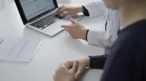 A person in a white lab coat with a stethoscope around their neck types on a laptop while another person looks on.