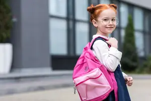 A young girl with red hair is wearing a white long-sleeve shirt and a blue skirt, carrying a pink backpack, and smiling in front of a blurry background with a glass wall and plants.
