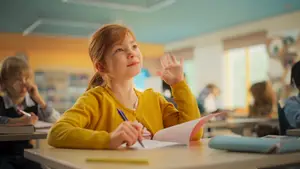A girl sitting at a desk in a classroom is holding a pen and a book and raising her hand