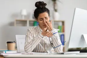 Woman sitting at a desk, rubbing her forehead with her hand while holding a pair of glasses in the other.