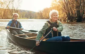 Two older adults are paddling a canoe on a river during the daytime, and they seem to be enjoying the view.