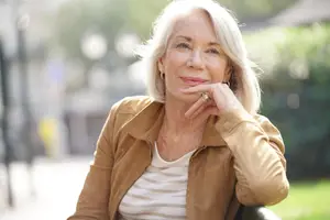An older woman with white hair is sitting on a bench in a park, smiling, and has her hand on her chin.