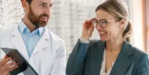 An adult woman trying on glasses in front of an optometrist wearing a white coat