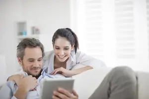 A man and woman sitting on a couch with the woman looking at a tablet while the man holds it.