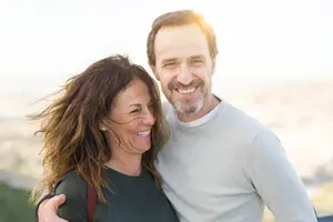 A smiling man and woman are standing close to each other with their arms around each other.