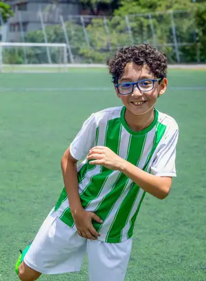 A boy in a green and white striped shirt and blue glasses poses on a soccer field.