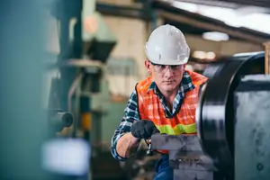 A man in a factory wearing a white hard hat, orange reflective vest, and black gloves is working on a large metal machine.