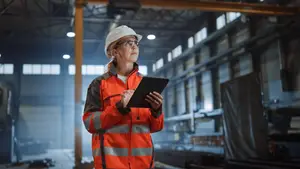 An adult female in a reflective safety jacket and hard hat is standing in a factory and looking at a tablet.