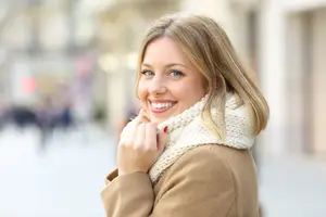 A woman wearing a brown coat and a white scarf is smiling and looking forward on a sunny day.