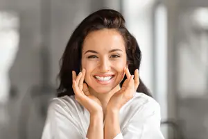 A woman is smiling and posing for a picture while holding her cheeks with both hands.
