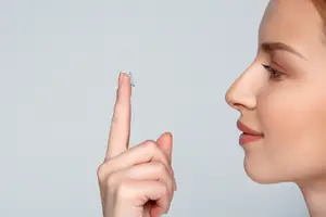 A woman is holding a blue contact lens in her finger and showing it to the camera.