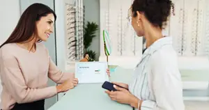 A woman in a white shirt is holding a phone and looking at a tablet while a woman in a pink shirt smiles and holds a tablet in an office. Behind them is a plant and a mirror.
