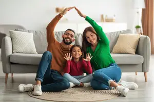 A family of three is sitting on a couch in a living room, smiling and posing for a photo. The man is making a peace sign with his hands, and the woman is making a peace sign with her hand while holding the girl's hand. The girl is smiling and showing a peace sign with both hands. The couch is gray and has white pillows, and there is a carpet on the floor. Behind the couch is a white shelf with a potted plant and a lamp.