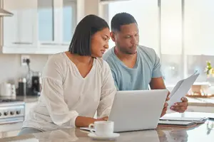 A man and a woman are sitting at a table in a kitchen, working on a laptop with papers on it.