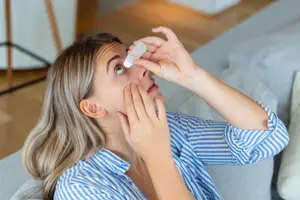 A woman with a blue striped shirt on is sitting on a couch and looking up with her hand on her face while holding a bottle of eye drops.