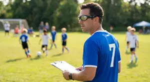 A man wearing a blue soccer jersey is standing on a soccer field and looking at a clipboard.