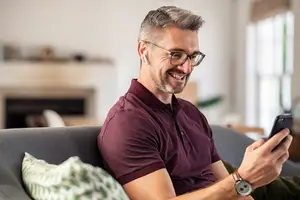 A smiling man with glasses and a watch is sitting on a couch, holding a cell phone in his right hand, and probably talking to someone.