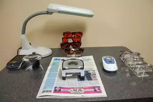Table with various items including a lamp, sunglasses, a newspaper, a magnifying glass, and a remote control.