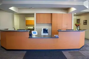 An interior of an office with a reception desk, a computer monitor, a printer, a monitor, and a vase with flowers on a wooden counter with a black carpet in front of it.